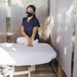 Female Therapist Wearing Mask Standing By Massage Table In Spa Stock Photo