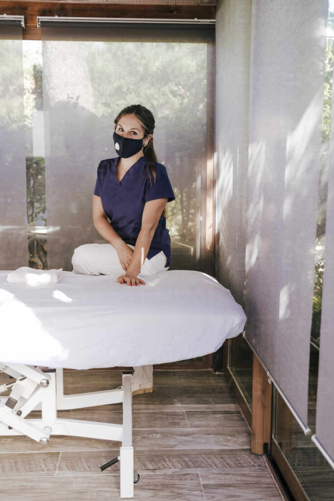 Female Therapist Wearing Mask Standing By Massage Table In Spa Stock Photo